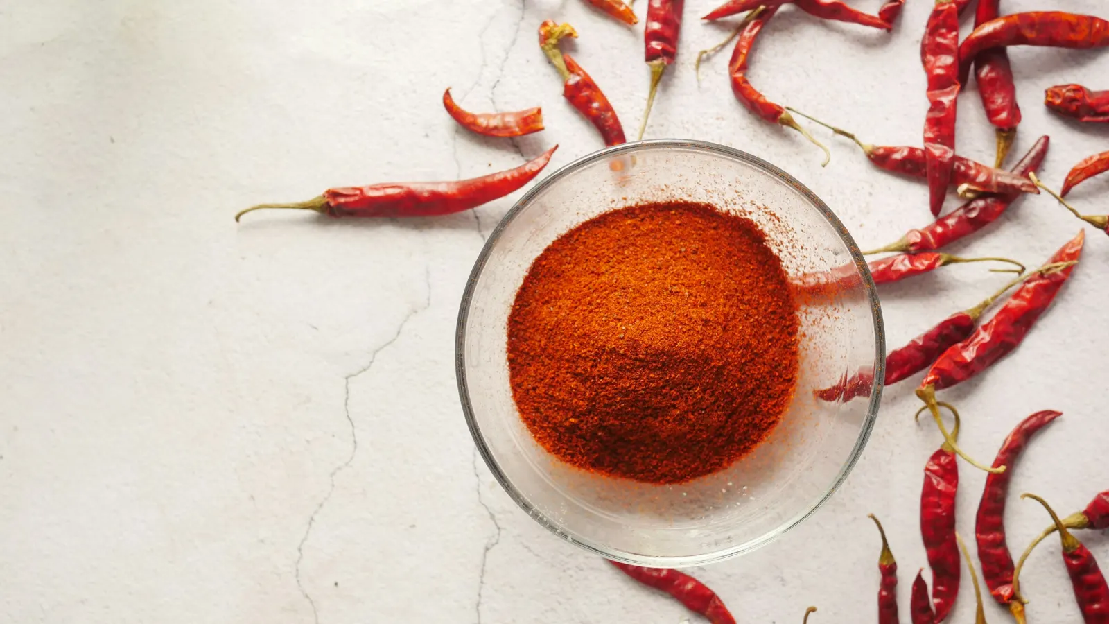 Glass bowl of ground red chilli surrounded by dried red chillies on a pale surface