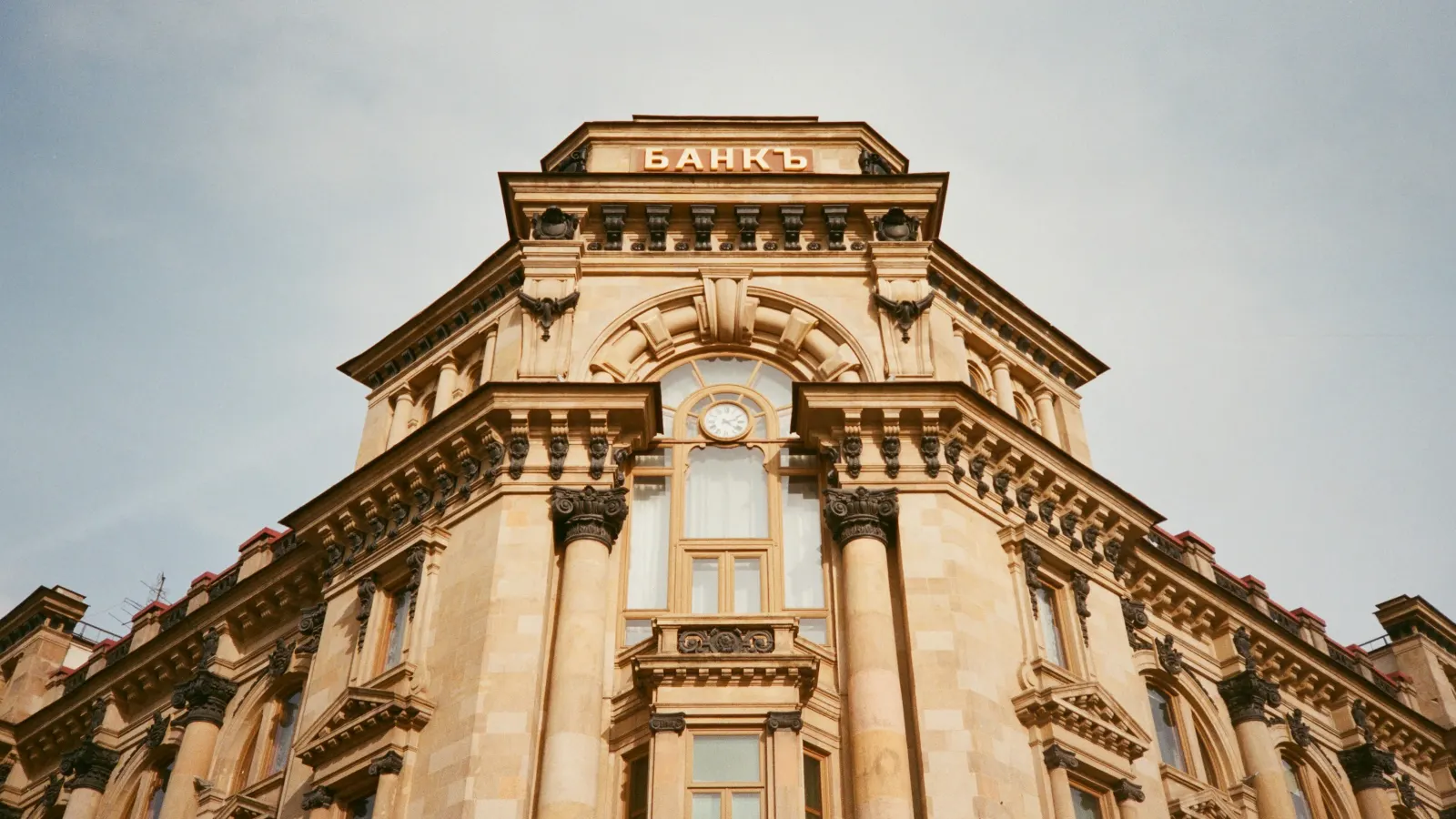 Ornate sandstone corner of a historic bank building with a clock and the pre-reform Russian word for 'bank' above the upper storey