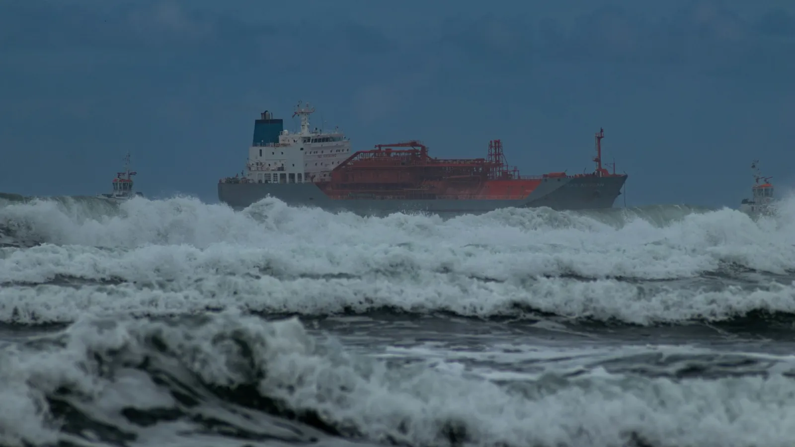 Tanker ship riding out breaking storm waves close to shore under a dark sky