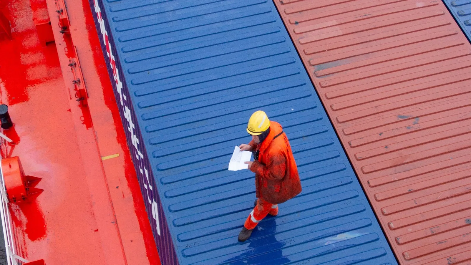 Worker in a hard hat and high-visibility coat reading a paper list while walking across container hatch covers on a ship's deck