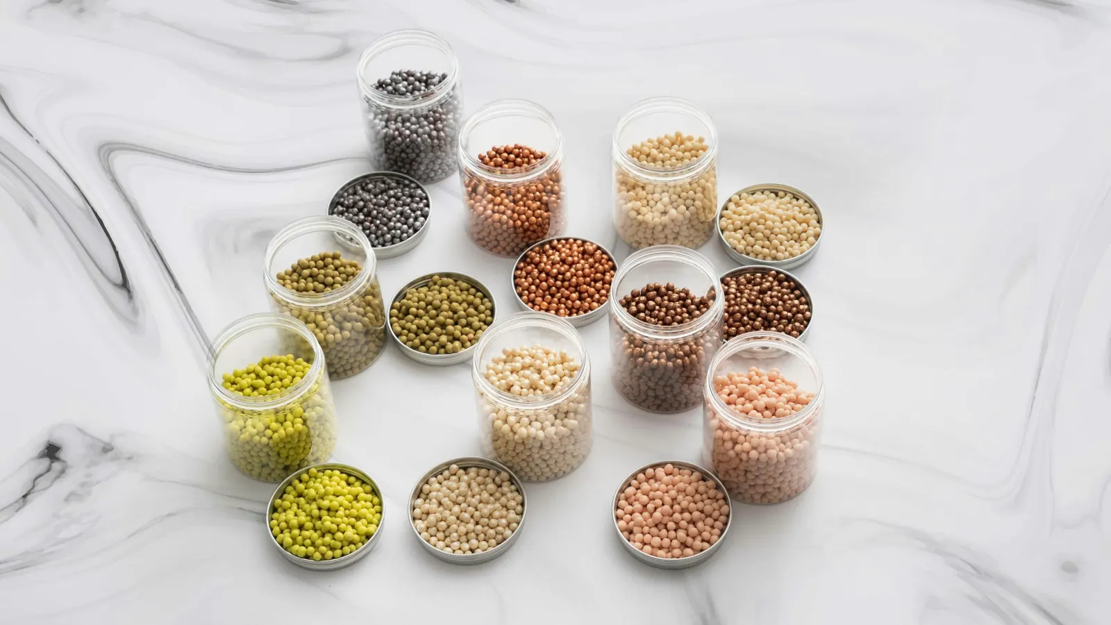 Open clear sample jars, each holding a different batch of small round pellets, beside their lids on a marble surface