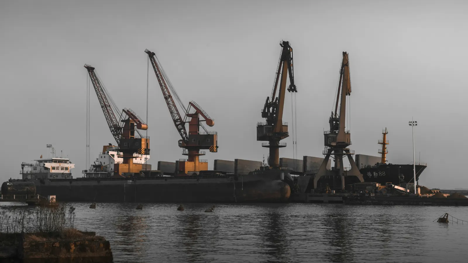 Port cranes loading a bulk cargo ship moored at a quay under a grey sky