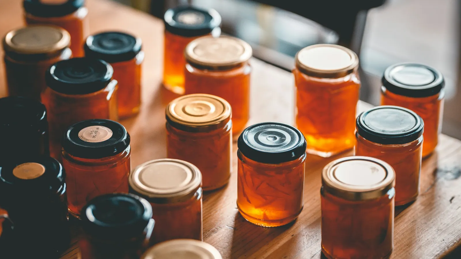 Unlabelled glass jars of the same orange preserve on a wooden table, some with gold lids and some with black lids