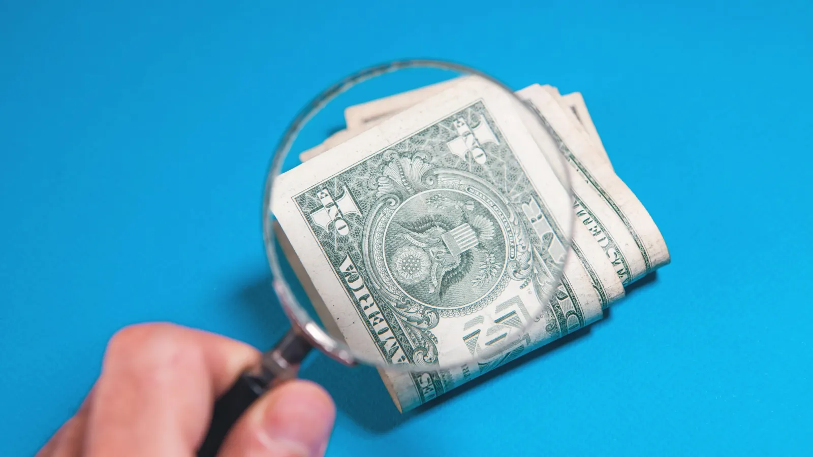 Magnifying glass held over folded US one-dollar bills on a blue background