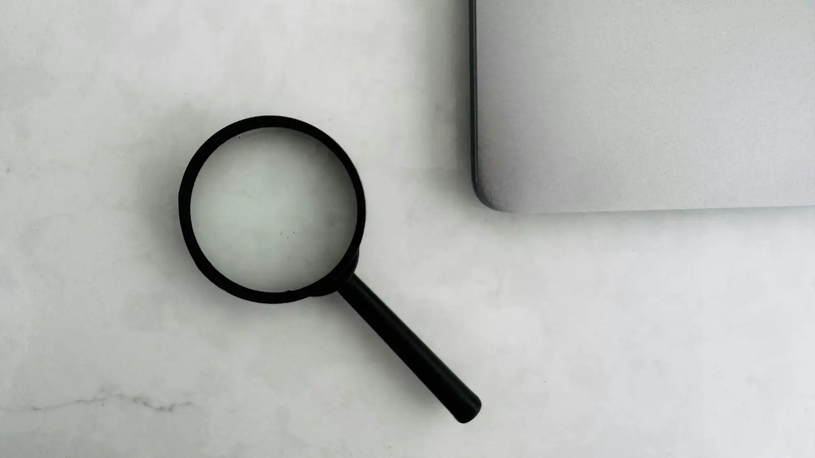 Magnifying glass resting beside the corner of a laptop on a white marble surface