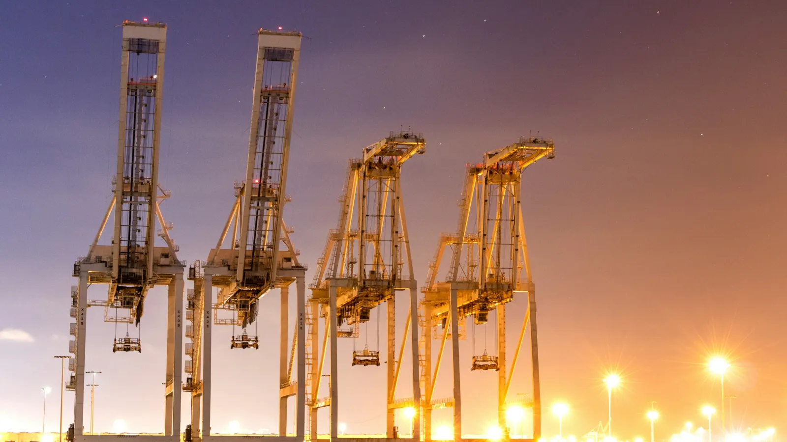 Four ship-to-shore container cranes lit against a dusk sky at a container terminal