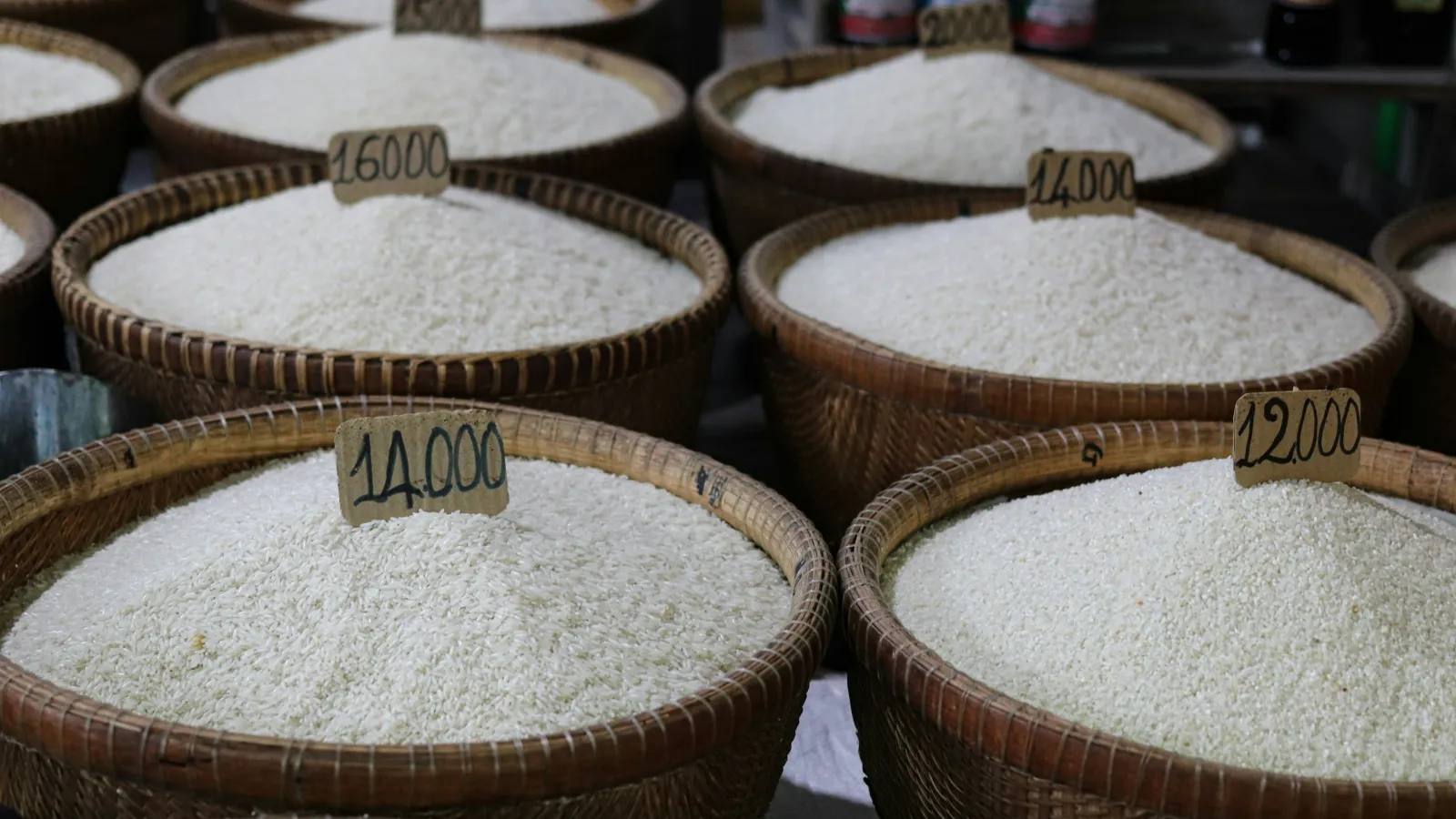 Several woven baskets of white rice from different lots, each with a handwritten price tag, at a wholesale market